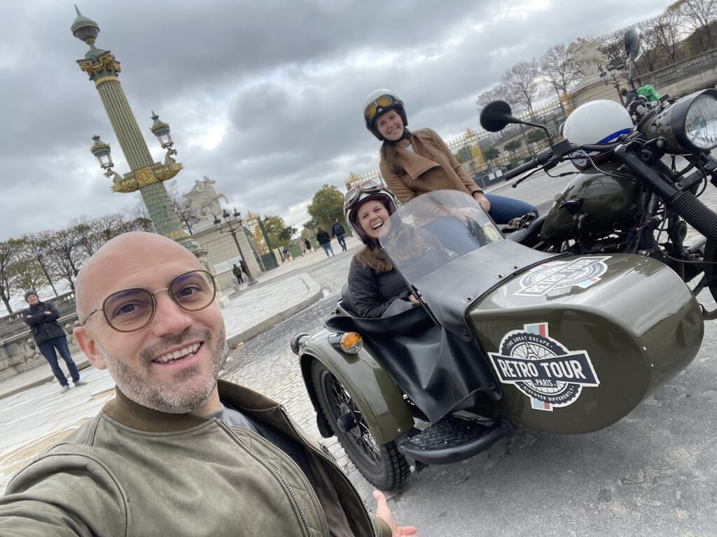 A joyful selfie of a man with a woman sitting in a sidecar and another woman on the motorcycle, with the ornate Place de la Concorde column in the background, epitomizing a private vintage motorcycle tour in Paris, France.