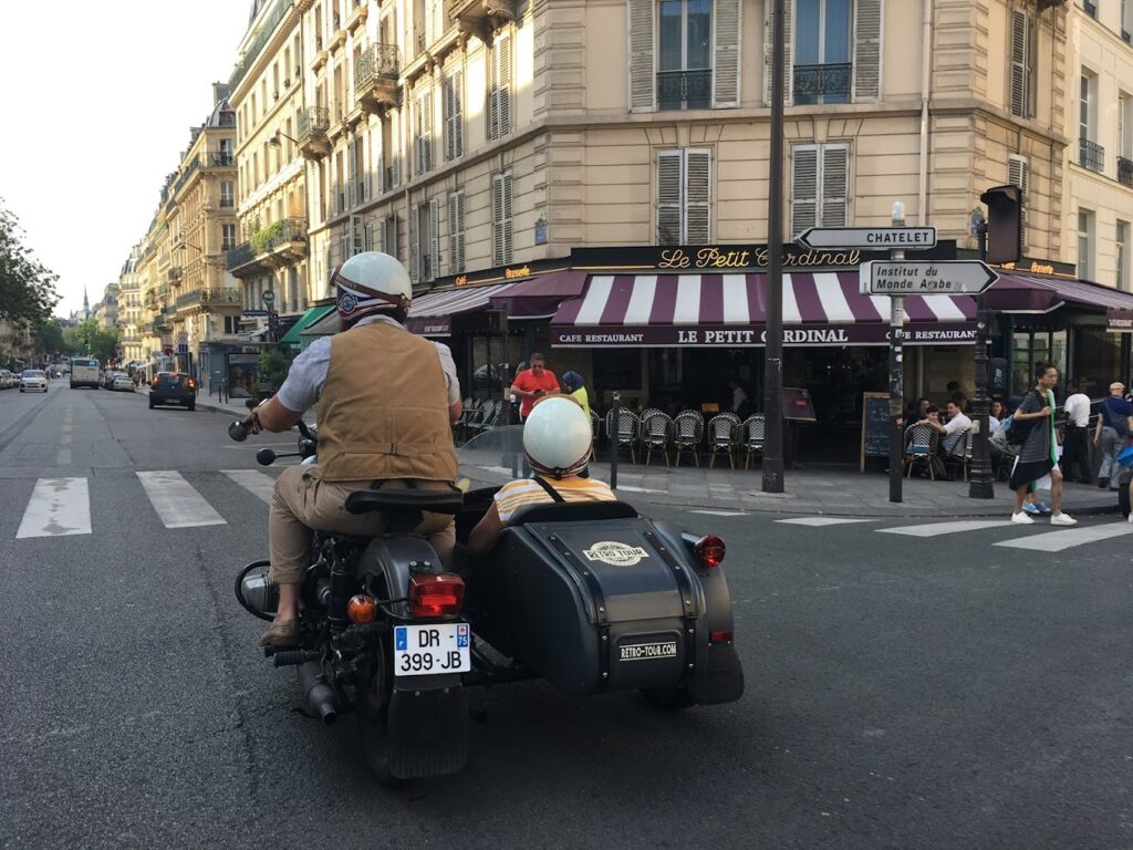 A passenger in a vintage sidecar motorcycle enjoys a ride through a bustling Parisian street, passing by the charming café 'Le Petit Cardinal,' a classic Paris experience for motorcycle tours in France.