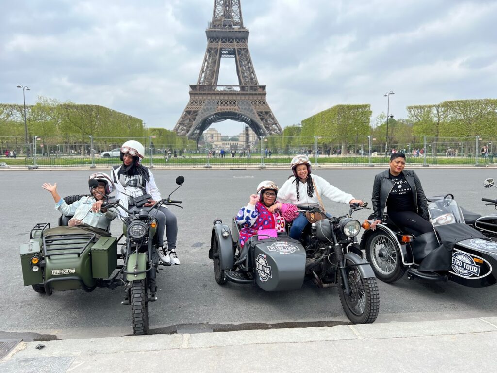A group of excited tourists on sidecar motorcycles waving and posing in front of the Eiffel Tower, a memorable highlight of their motorcycle tour in Paris, France.