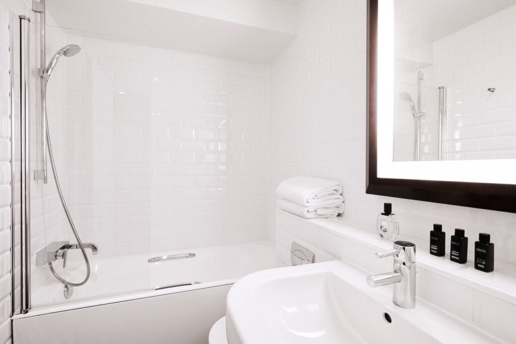 Modern bathroom with pristine white subway tiles, a sleek bathtub with shower attachment, a large mirror with backlighting, and a neat selection of luxury toiletries, reflecting the clean and sophisticated design typical of upscale Paris hotels.