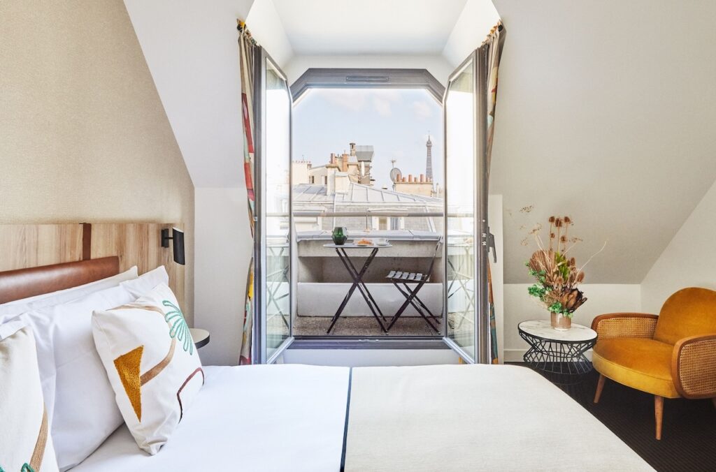 Chic hotel room with a crisp, white bed facing a large bay window that opens to a quaint balcony with a view of the Eiffel Tower, accompanied by vibrant decorative touches and a modern, cozy armchair, epitomizing the stylish comfort of Parisian hotels with iconic views.