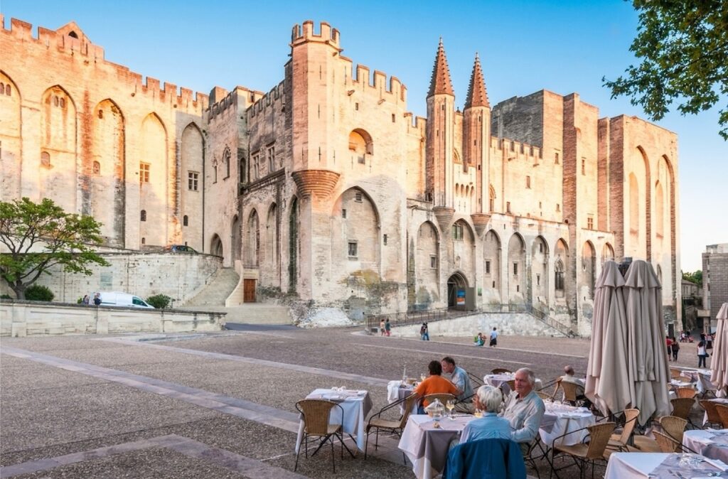 Diners at an outdoor restaurant enjoying a meal with a stunning view of the Pope's Palace in Avignon, a historic gem among the best cities in the south of France, bathed in the warm glow of sunset.