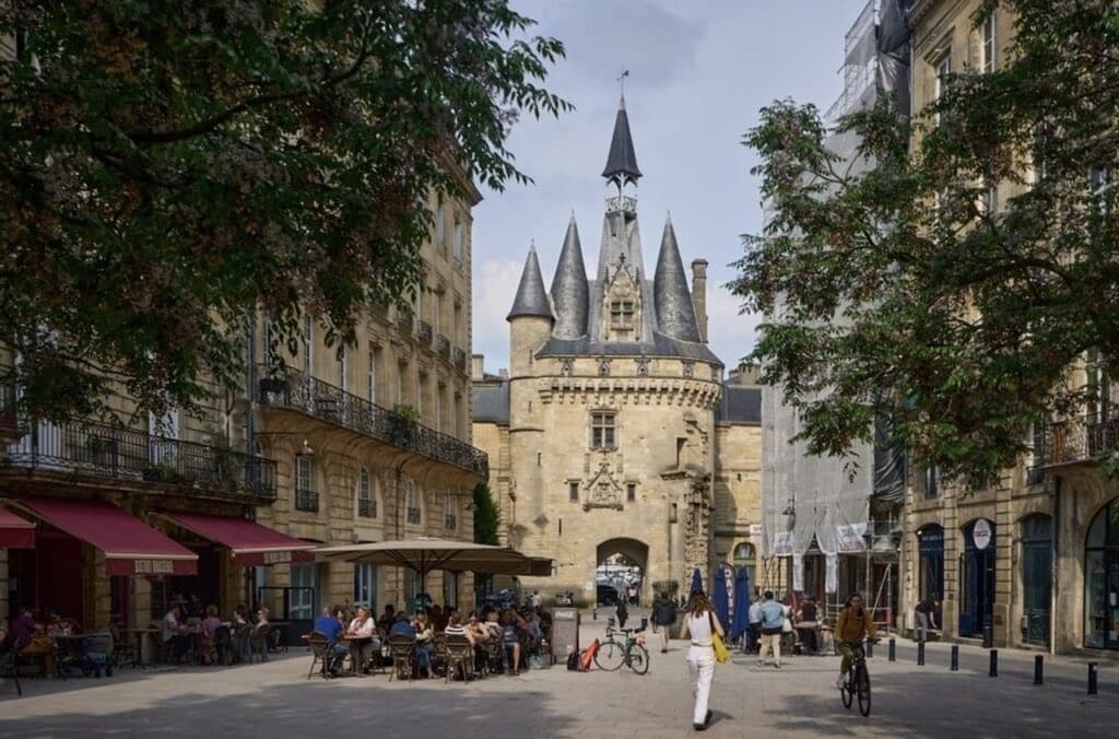 Visitors dining at an outdoor café near Porte Cailhau in Bordeaux, enjoying a leisurely moment during their one day in Bordeaux. The iconic medieval gate stands in the background, its gothic architecture and turrets a stark contrast to the surrounding modern city life.