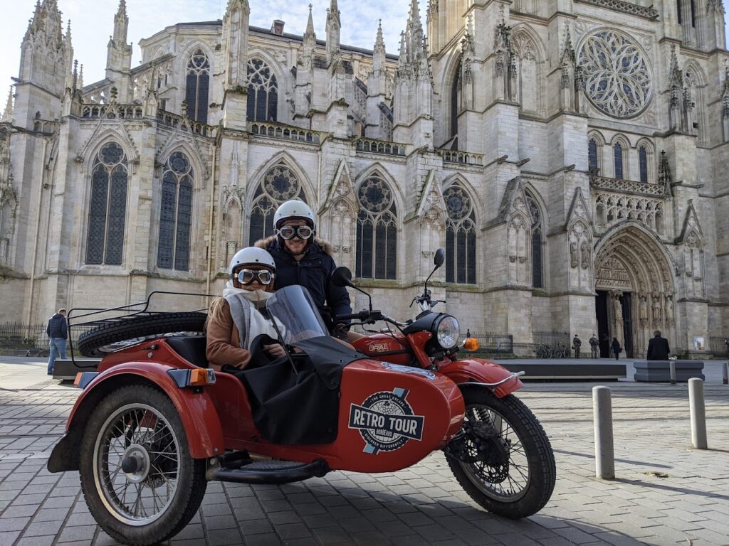 Two tourists on a red sidecar motorcycle in front of the intricate Gothic architecture of Bordeaux Cathedral, exploring the historic city on a unique motorcycle tour in France.