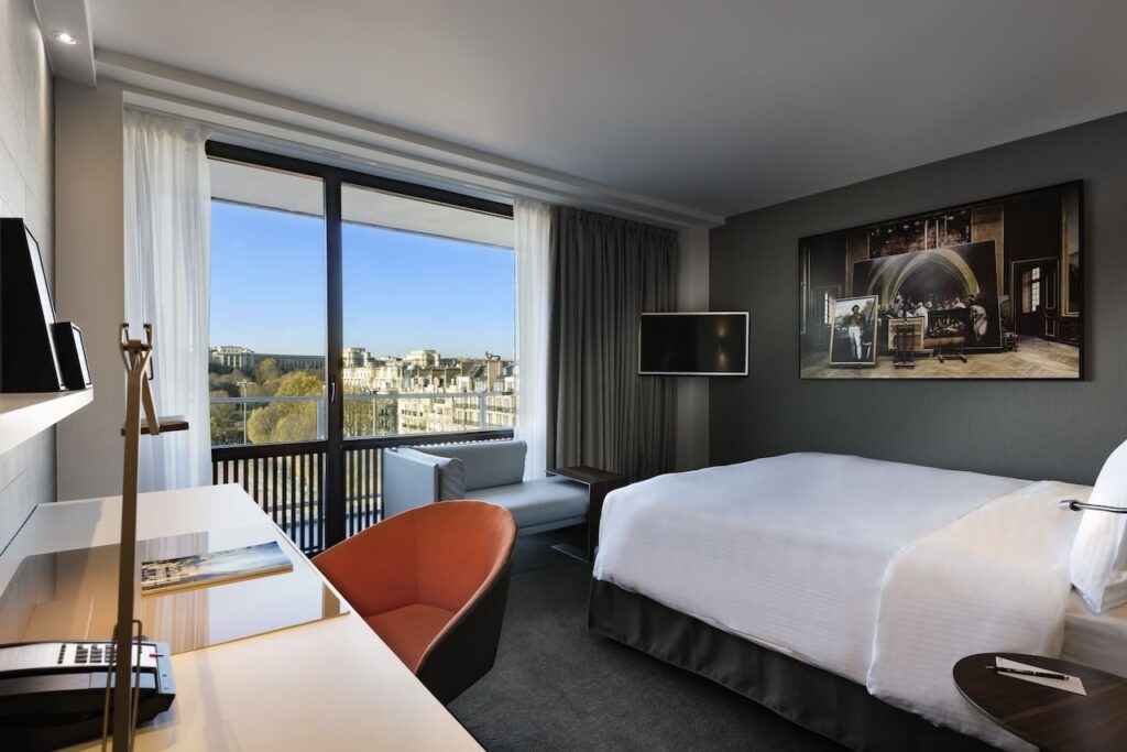 Stylish and contemporary hotel room at the Pullman Paris Tour Eiffel featuring a comfortable bed with crisp white linens, a modern artwork of a historic scene above the bed, sleek furnishings, and a large window offering a breathtaking view of the Parisian skyline.