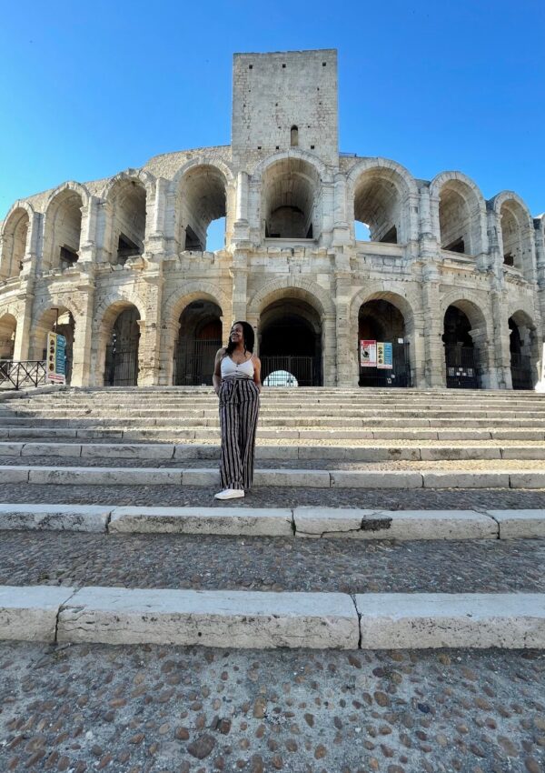 A woman stands on the steps of the Arles Roman Amphitheatre, looking up in admiration at the ancient stone arches against a clear sky, an essential experience for any itinerary on the best things to do in Arles.