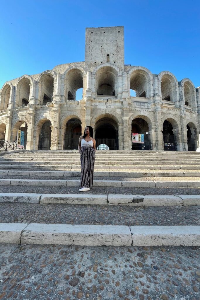 A woman stands on the steps of the ancient Roman Amphitheatre in Arles, a landmark reflecting the rich history of one of the best cities in the south of France, under a clear blue sky.