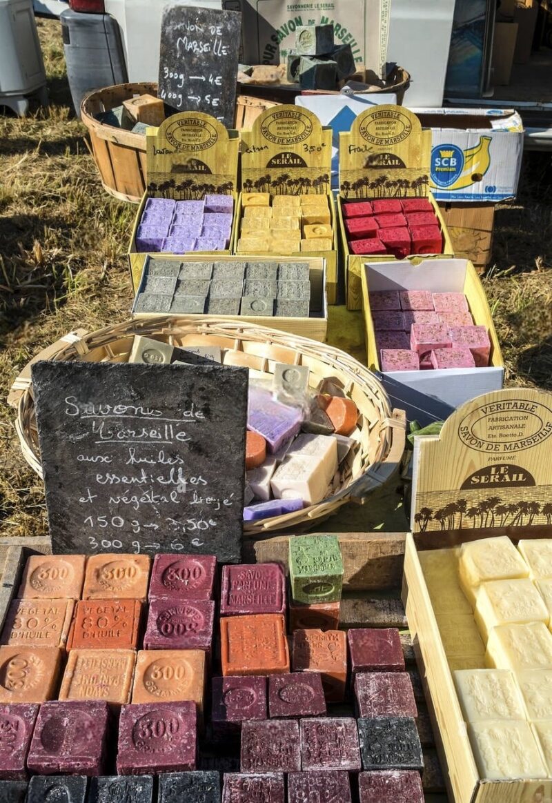 Outdoor market stall featuring a variety of handmade Marseille soaps with essential oils, displayed with pricing on a chalkboard sign, ideal for those seeking authentic Marseille souvenirs.