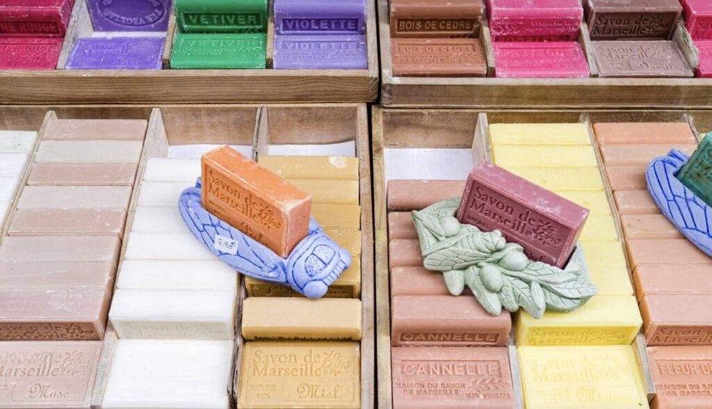Assortment of colorful traditional Marseille soap bars arranged in wooden compartments, with labels such as 'Vetiver,' 'Violette,' and 'Bois de Cedre,' perfect as Marseille souvenirs.