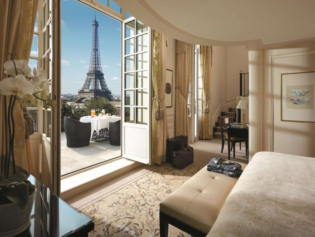 Luxurious hotel room with opulent decor, opening onto a balcony with a table set for two, offering a breathtaking direct view of the Eiffel Tower in a serene Parisian morning, a quintessential experience for guests at the Shangri-La Paris, known for its proximity to the iconic landmark.