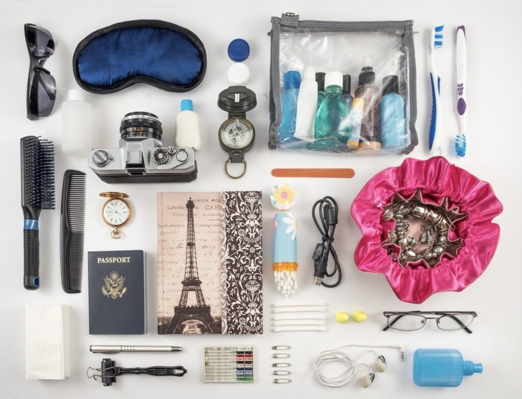 A flat lay of travel essentials including a passport, camera, toiletries bag, eyemask, sunglasses, hairbrush, notebook with Eiffel Tower, and various chargers—a meticulously organized packing list for France, ensuring preparedness for every aspect of travel.