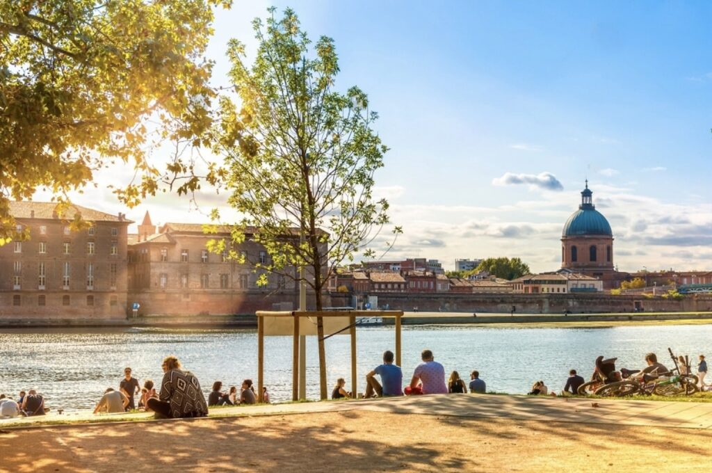 A serene riverside scene in Toulouse, one of the best cities in the south of France, with locals enjoying the sunshine on the grassy bank, facing historical red-brick buildings and the iconic dome of La Grave.