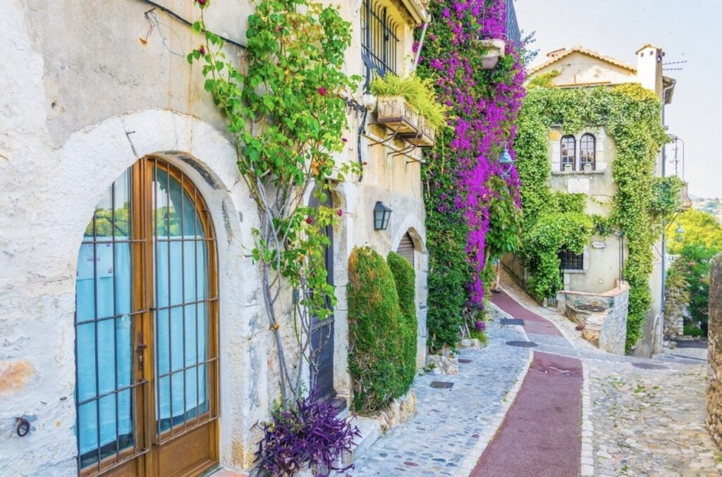 Charming cobblestone street in Saint-Paul-de-Vence, showcasing the best of southern France's architecture with ivy-covered walls and vibrant bougainvillea framing a serene view of the azure sea.