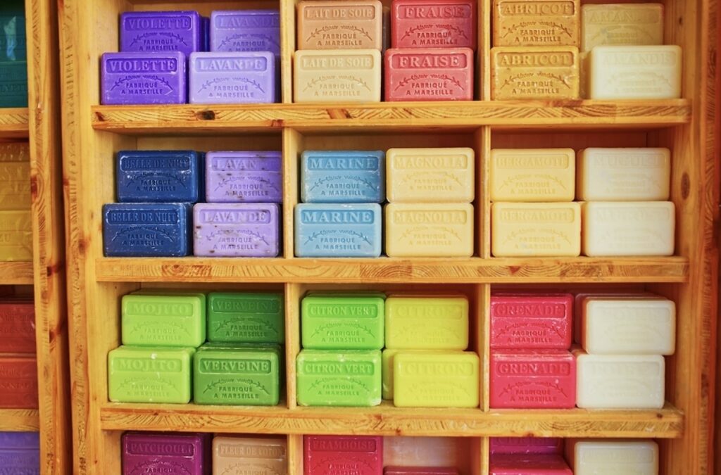 Vibrantly colored Marseille soap bars neatly organized on wooden shelves, with a range of scents labeled in French like 'Violette,' 'Lavande,' 'Marine,' and 'Mojito,' capturing the essence of Marseille souvenirs.