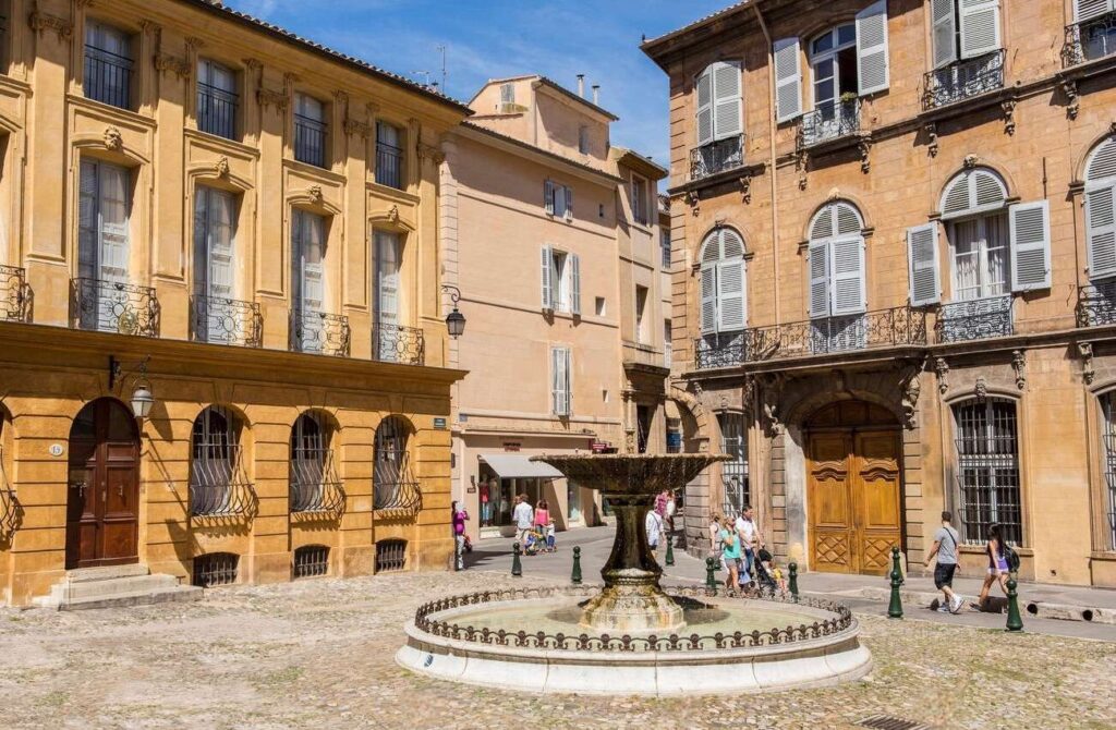 The quaint charm of Aix-en-Provence with pedestrians strolling by a classic fountain in a cobblestoned square, flanked by traditional ochre buildings with intricate ironwork, embodying the allure of the best cities in the South of France.