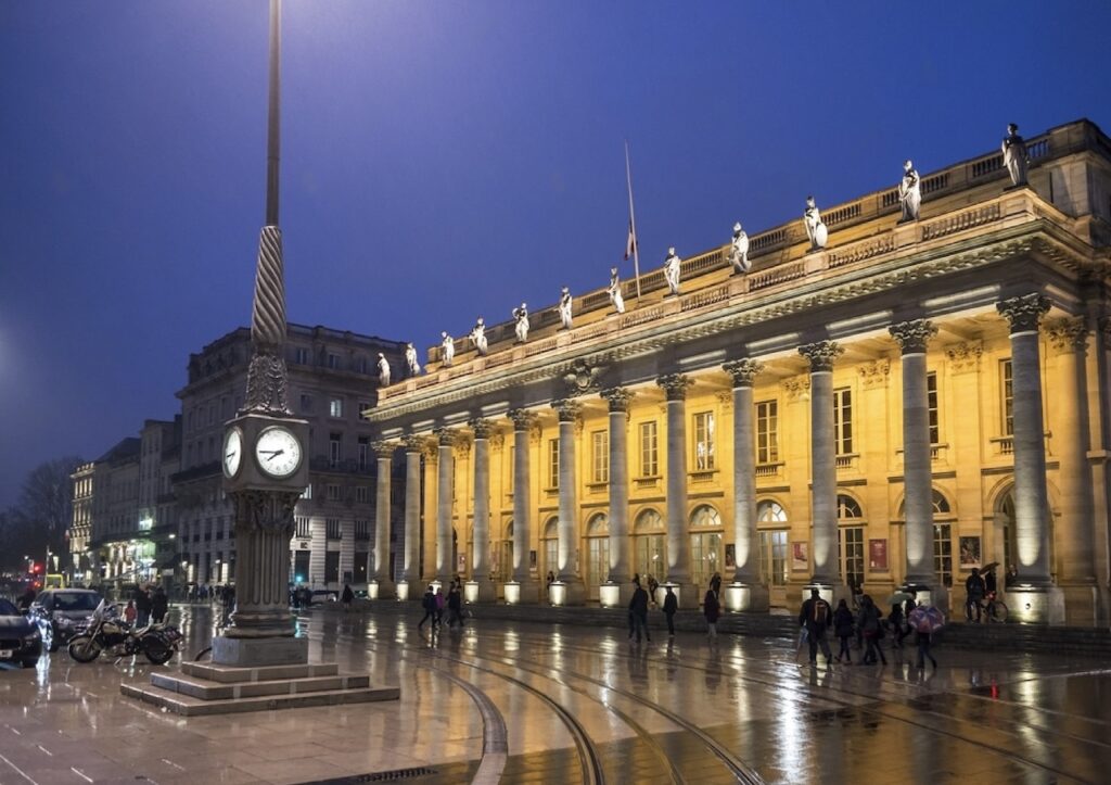 The Grand Théâtre de Bordeaux stands majestically illuminated at night, a beacon for those exploring the city in one day in Bordeaux. The wet cobblestone streets reflect its neoclassical architecture and statues, with passersby and the ornate street clock adding to the evening's ambiance.