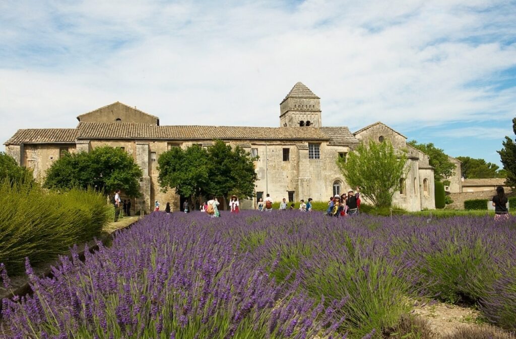 Visitors wander near an ancient stone abbey surrounded by vibrant lavender fields under a sunny sky, a scene capturing the rustic charm of the best cities in the South of France.