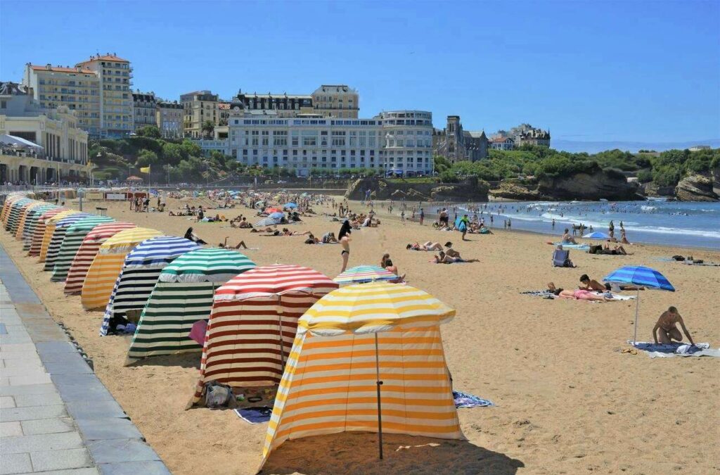 The sandy beach of Biarritz lined with a colorful array of striped beach tents, against a backdrop of historic buildings and azure waters, showcasing the laid-back coastal lifestyle in the best cities in the South of France.