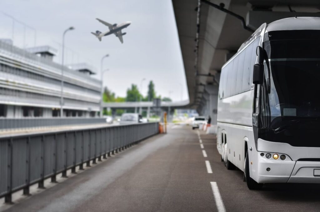 A modern shuttle bus waits on the tarmac of an airport with an airplane taking off in the blurred background, a common transport option for 'Getting From Beauvais Airport to Disneyland Paris' for both convenience and comfort.