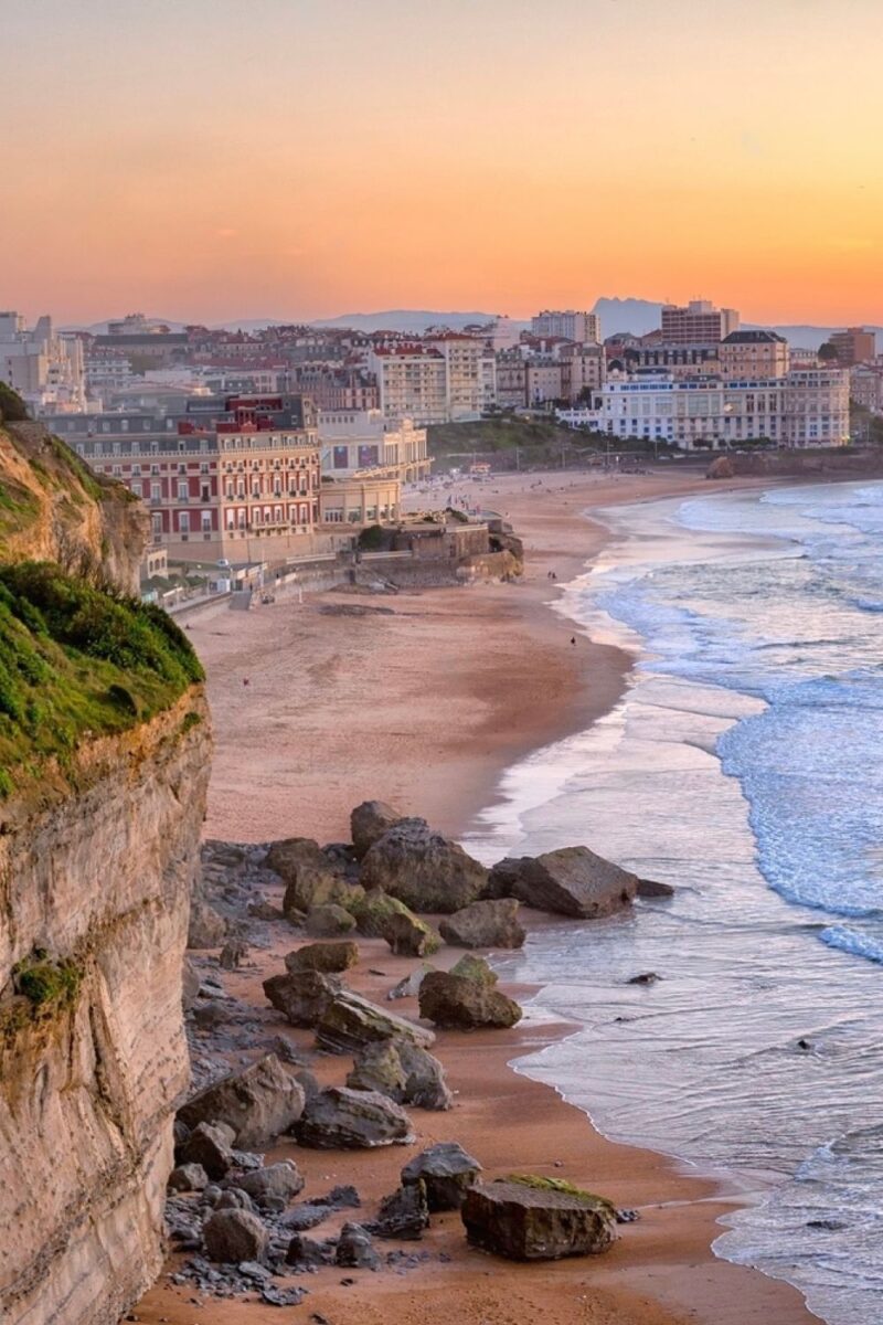 Sunset over Biarritz beach, with the orange hues of the setting sun casting a warm glow on the cityscape and sandy shores, a serene moment in one of the best cities in the South of France.