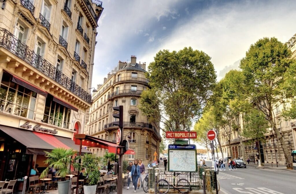 A lively Parisian corner in the 6th arrondissement featuring the iconic Art Nouveau Métropolitain sign of the Assemblée Nationale station, bustling sidewalk cafés, and traditional Haussmannian architecture, illustrating the area's appeal as one of the best arrondissements to stay in Paris.