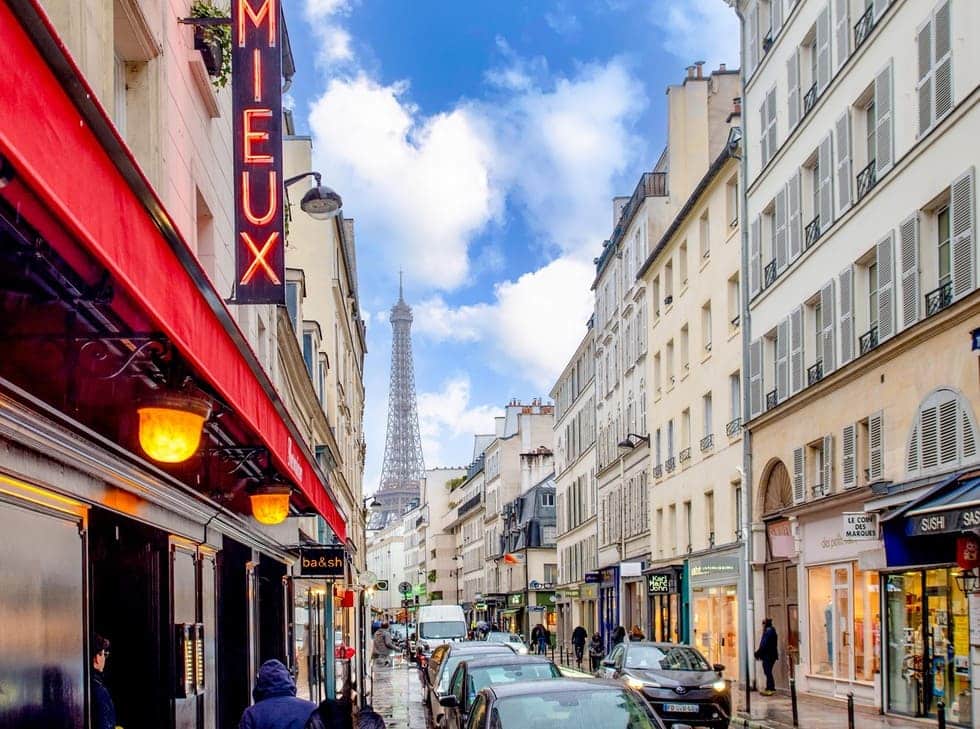 The charming streets of the best arrondissement to stay in Paris on a clear day, with the iconic Eiffel Tower in the distance and vibrant storefronts lining the road, as pedestrians and cars share the bustling thoroughfare.