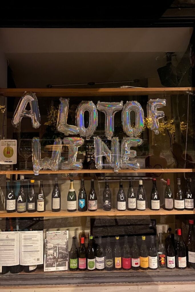 A whimsical display of metallic balloon letters spelling 'A LOT OF WINE' hovering above an array of diverse wine bottles on wooden shelves, indicative of the variety and charm found at the best wine bars in Paris.