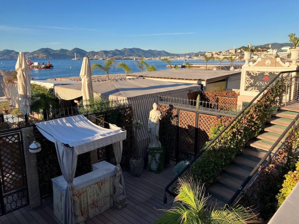 Early morning at La Plage du Festival in Cannes, with the club's elegant wooden deck, lush potted plants, and neatly arranged sun loungers awaiting guests. The calm sea and the silhouette of the Esterel Mountains in the distance offer a tranquil seaside escape, characteristic of Cannes beach clubs.