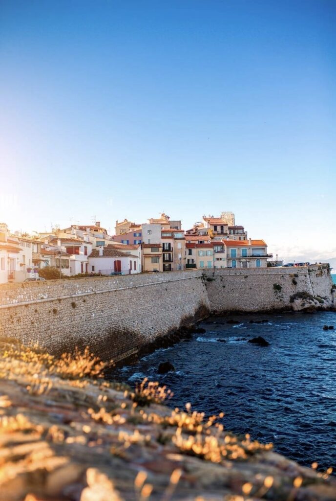 Golden sunlight bathes the historic sea wall and traditional pastel-colored houses of Antibes, a charming coastal town renowned as one of the must-visit the French Riviera cities. The calm Mediterranean Sea gently laps against the rugged shoreline, capturing the essence of this picturesque seaside escape.