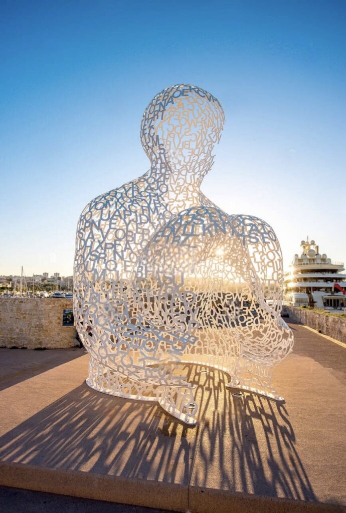 An artistic sculpture made of intricate letter-shaped cutouts forming the silhouette of a seated person against a bright blue sky, located in Antibes, one of the must-visit French Riviera cities. The sculpture casts a striking shadow on the ground, emphasizing its unique design and the serene Mediterranean backdrop.