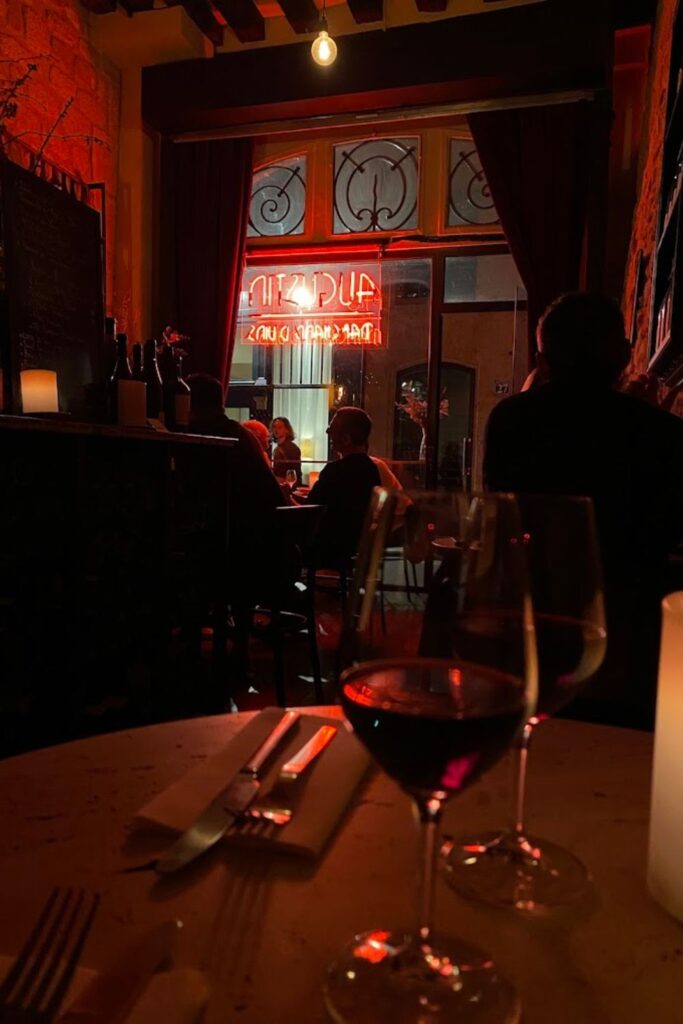 Intimate ambiance at one of the best wine bars in Paris, showcasing a candlelit table set for two with glasses of red wine, against a backdrop of patrons and a glowing neon sign in reverse reflection through a window.