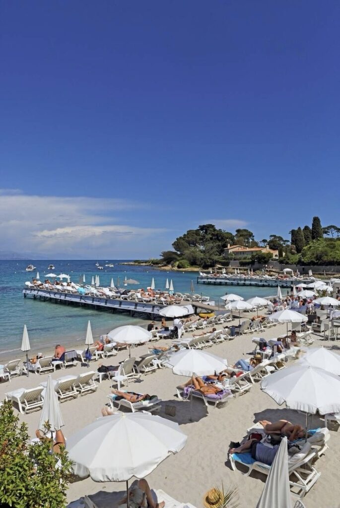 Visitors relaxing under white umbrellas on sandy shores at one of the best beach clubs in Antibes, with a wooden pier extending into the serene blue waters of the Mediterranean Sea, against a backdrop of lush greenery and clear skies.