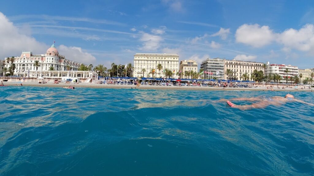 Swimmer's perspective of the Mediterranean with a view of the distinguished architecture of Beach Clubs in Nice, showcasing a promenade lined with parasols and bathers, under a bright blue sky.