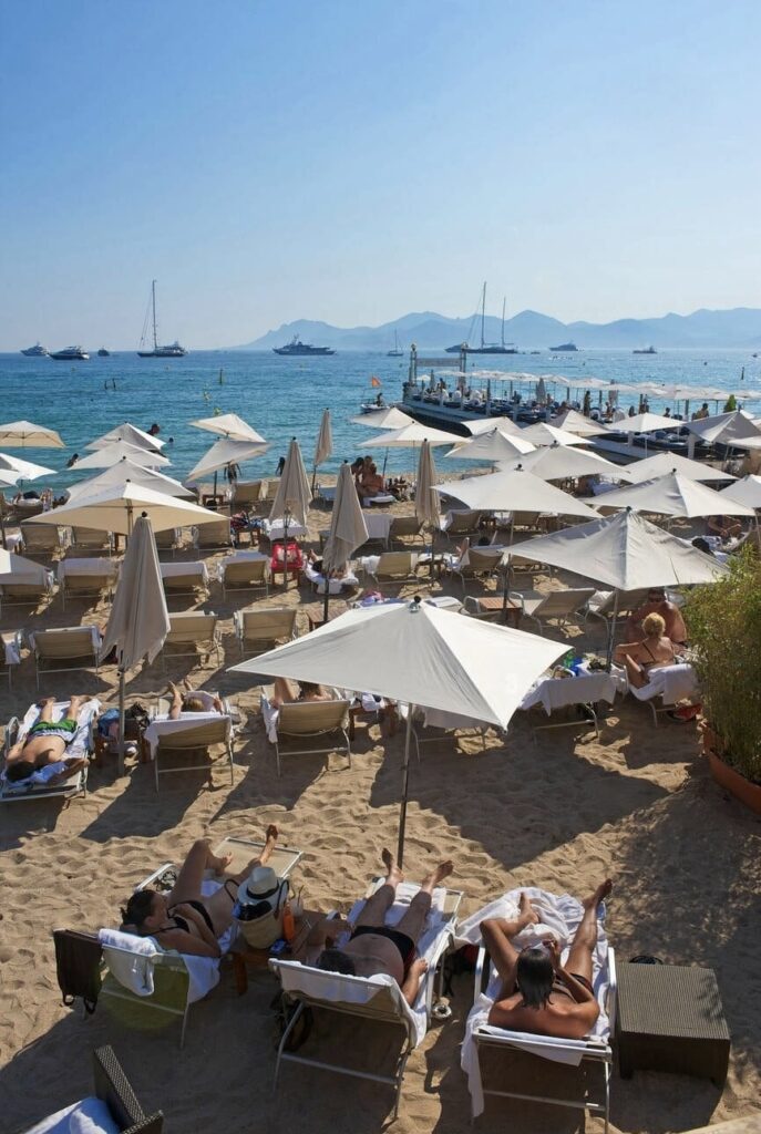 Visitors lounging on sunbeds under white umbrellas on a sandy beach in Cannes, a premier must-visit French Riviera cities. The tranquil blue sea dotted with yachts and sailboats under a clear sky encapsulates the luxurious and relaxing atmosphere of this famous seaside destination.