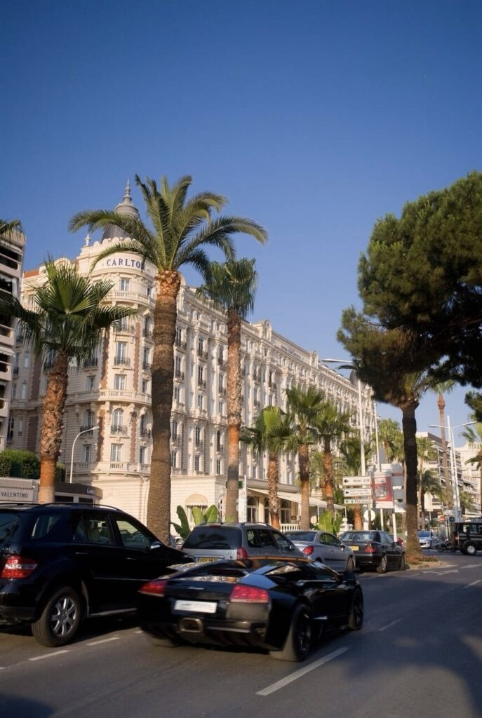 Luxury cars cruise by the iconic Hotel Carlton in Cannes, set against a backdrop of tall palm trees under a clear blue sky, showcasing the upscale ambiance of this must-visit French Riviera cities.