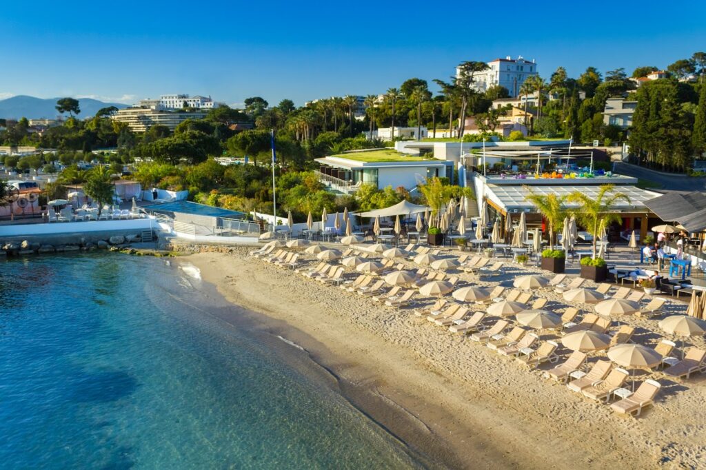 Cap d'Antibes Beach Hotel's exclusive beach clubs in Antibes, with rows of neatly aligned beige sun loungers and umbrellas on the sandy shore, overlooking the serene blue Mediterranean waters with lush greenery and elegant villas in the background.