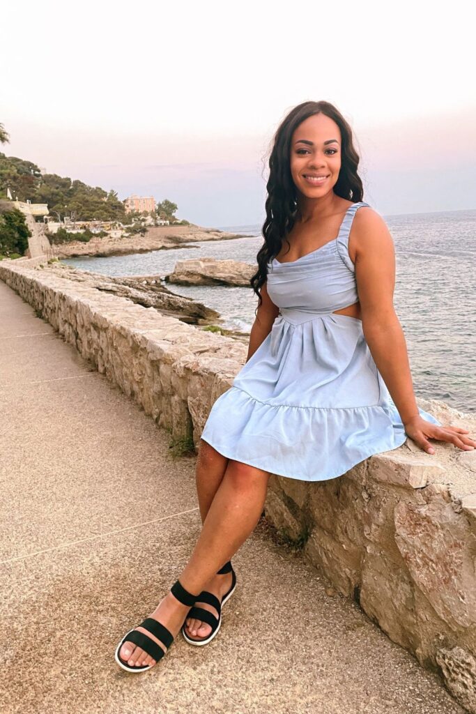 A woman in a light blue dress sits on a stone wall along a coastal promenade, with the serene Mediterranean Sea and a rocky shoreline in the background. This tranquil scene is characteristic of the leisurely pace and natural beauty found in the must-visit French Riviera cities.