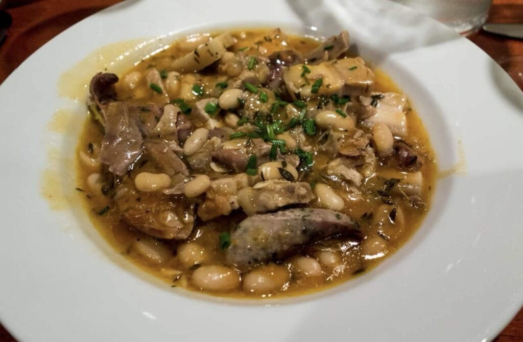 A sumptuous plate of Cassoulet, a beloved classic among famous French foods, with white beans, savory chunks of meat, and fresh herbs creating a rich, comforting stew, served on a white plate for a traditional French culinary experience.