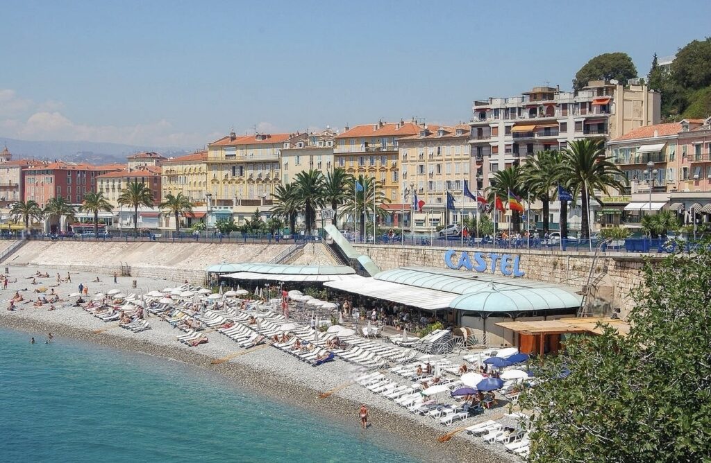 A bustling Castel Plage in Nice, France, with rows of sun loungers and parasols on a pebble beach, and the azure Mediterranean Sea beside it, capturing one of the best beach clubs in Nice.