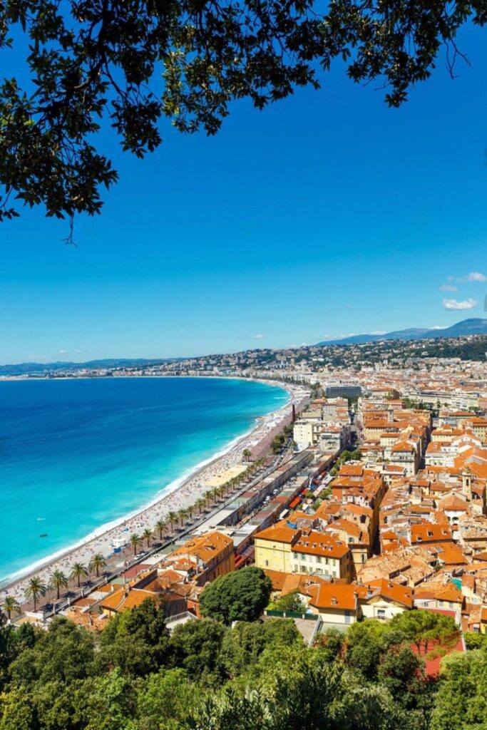 Panoramic view from Castle Hill overlooking the terracotta rooftops of Nice's old town, the famous Promenade des Anglais, and the stunning azure waters of the Bay of Angels, framed by lush green foliage. An essential highlight for any Nice travel guide, showcasing the city's picturesque blend of urban and natural beauty.