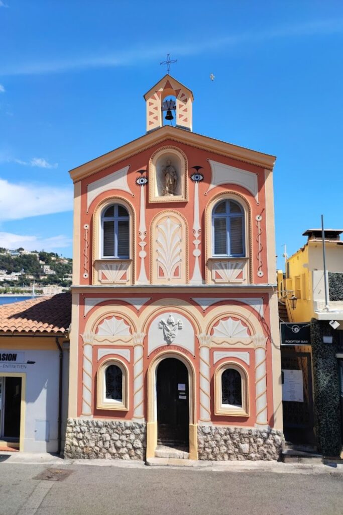 The distinctive Chapelle Saint-Pierre in Villefranche-sur-Mer, featuring ornate coral and cream facade, a small bell tower, and religious artwork above the entrance. A bright day accentuates the building's colorful details, a must-visit for those exploring things to do in Villefranche-sur-Mer.