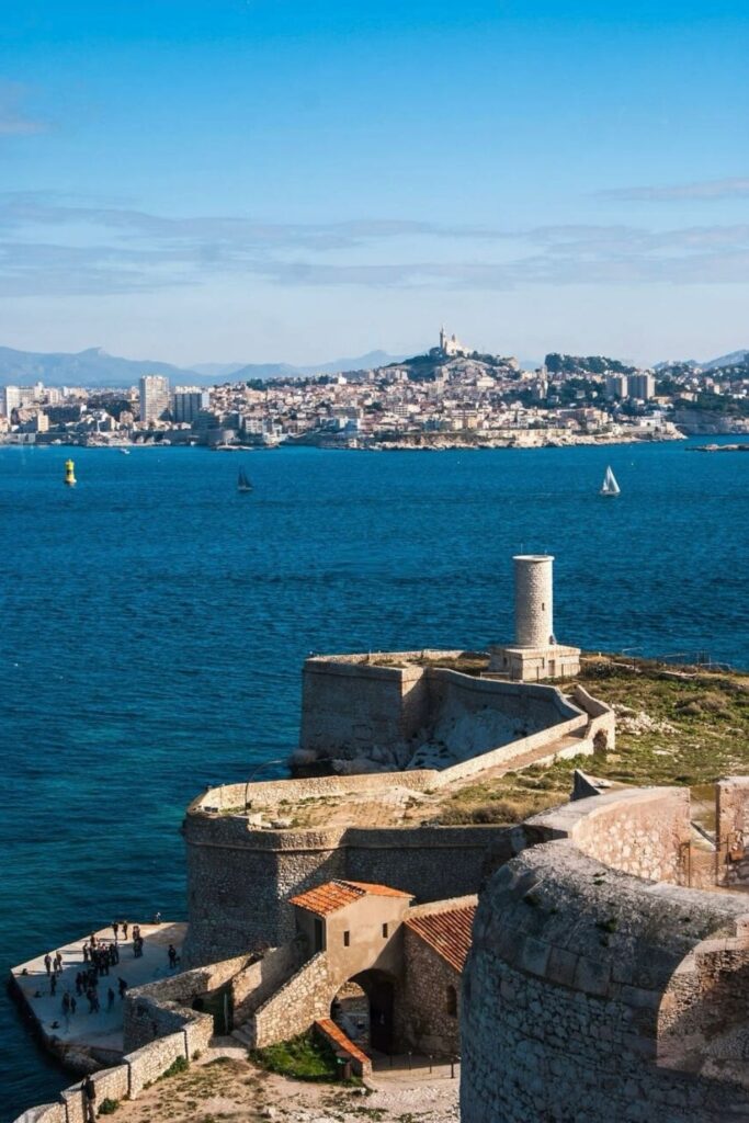 Historical Château d'If in the foreground overlooking the panoramic view of Marseille's coastline with sailing boats on the blue waters and the iconic Notre-Dame de la Garde in the distance, highlighting the must-visit places for things to do in Marseille.