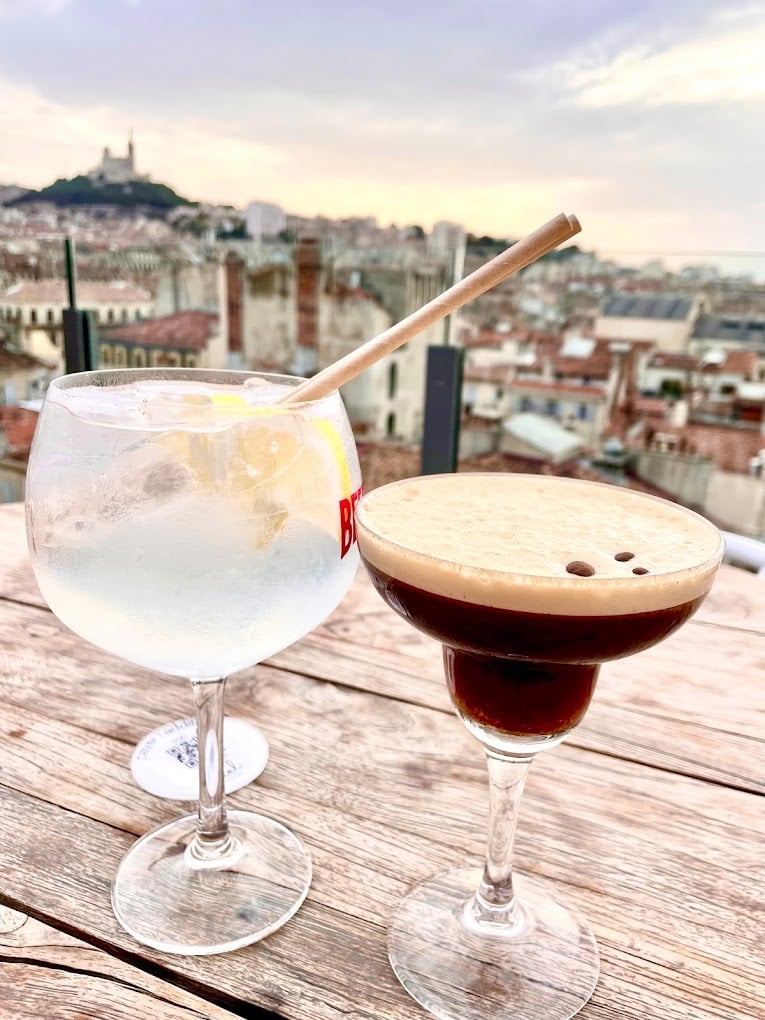 Two refreshing cocktails on a rooftop bar with a view of Marseille's cityscape and the iconic Notre-Dame de la Garde in the distance. The first cocktail is a gin and tonic with a bamboo straw, while the second is an espresso martini, both awaiting a toast to One Day in Marseille.