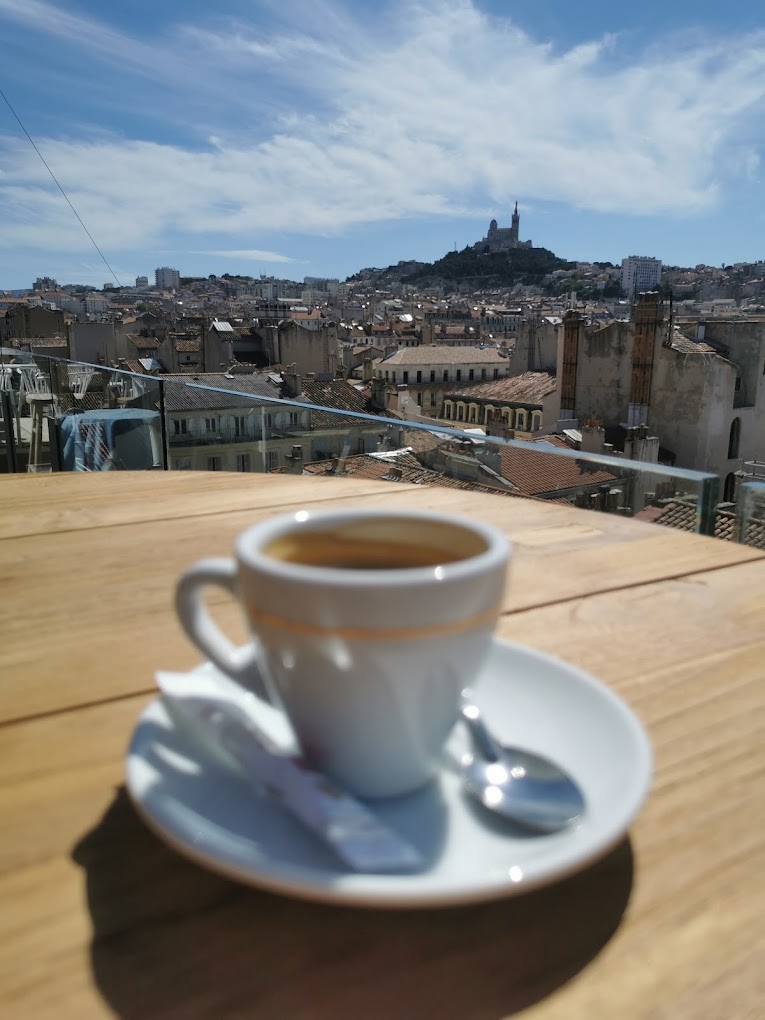 A cup of espresso sits elegantly on a saucer with a spoon, perched on a rooftop wooden table with a clear, sunny view of Marseille and the hilltop Notre-Dame de la Garde basilica in the background, capturing the essence of one day in Marseille.