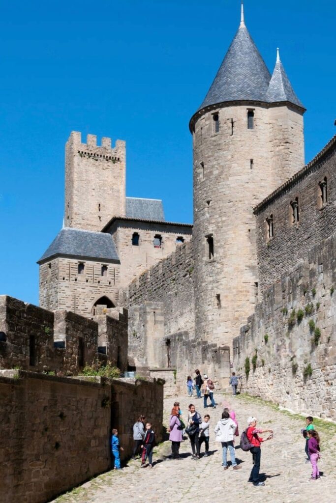 Tourists amble along the cobbled paths of the fortified medieval Cité de Carcassonne, with its imposing towers and ramparts under a clear blue sky, a popular historical day trips from Montpellier
