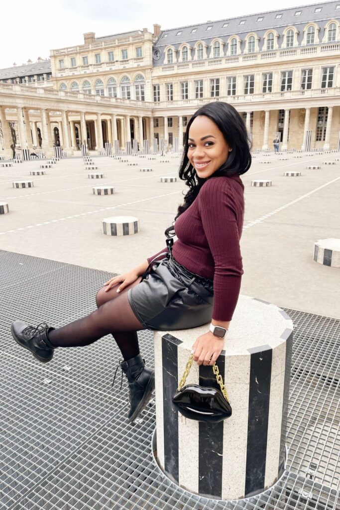 A radiant woman in a burgundy top and black leather skirt sits on one of the Colonnes de Buren in the Palais Royal, her smile reflecting the playfulness of this must-visit Paris instagram spots