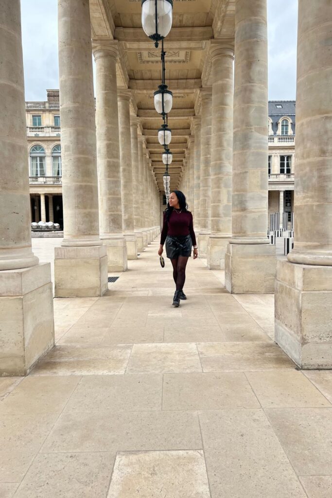 A woman walks between the grand stone columns of Palais-Royal, wearing a burgundy top and black skirt, with rows of elegant lanterns above, a popular Paris instagram spots