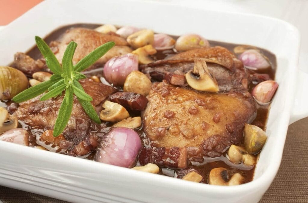 Coq au Vin, a signature dish among famous French foods, presented in a white casserole dish featuring braised chicken with shallots, mushrooms, and bacon, garnished with a sprig of fresh rosemary, epitomizing the rustic charm of French country cooking.