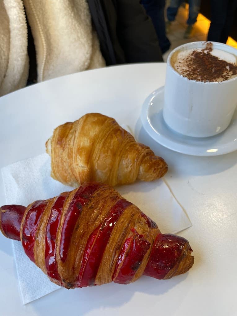 A cozy Parisian breakfast scene featuring famous French foods with a flaky, golden croissant and a raspberry-filled pain au chocolat on a white plate, alongside a cup of cappuccino with cocoa powder on top, evoking the warmth of a French café.