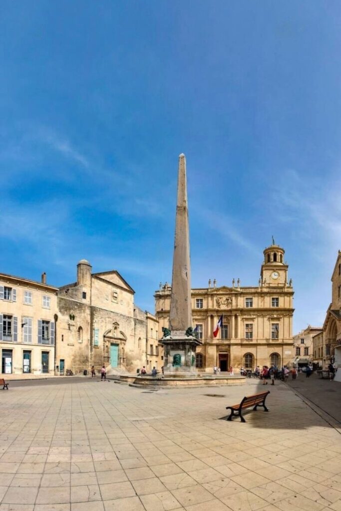 The bustling Place de la Republique in Arles with its iconic ancient obelisk, surrounded by historic architecture, including the city hall, under a clear blue sky—a quintessential stop for visitors seeking the best things to do in Arles.
